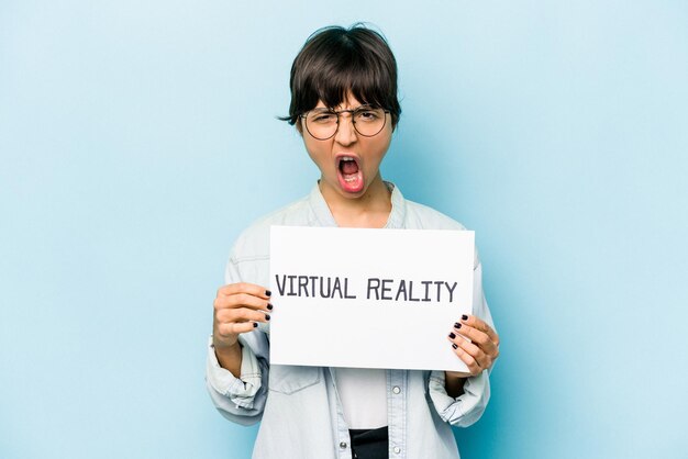 Młoda latynoska kobieta trzymająca tablicę wirtualnej rzeczywistości na niebieskim tle krzyczy bardzo zła i agresywna