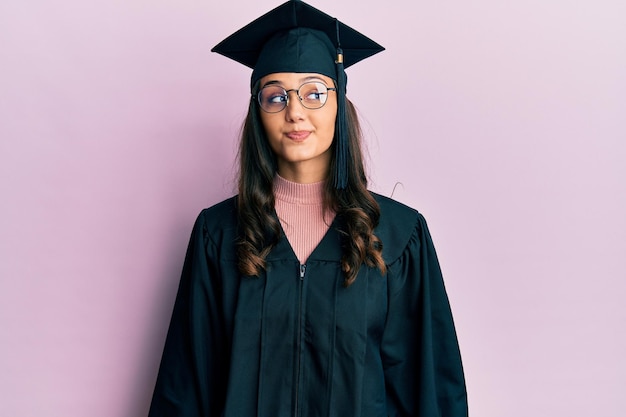 Młoda latynoska kobieta ubrana w czapkę dyplomową i szatę ceremonii uśmiecha się, patrząc w bok i odwracając wzrok, myśląc.