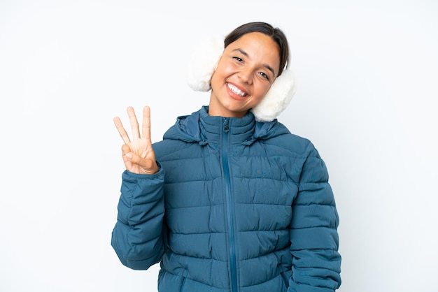 Młoda latynoska kobieta ubrana w nauszniki zimowe na białym tle szczęśliwa i licząca trzy palcami