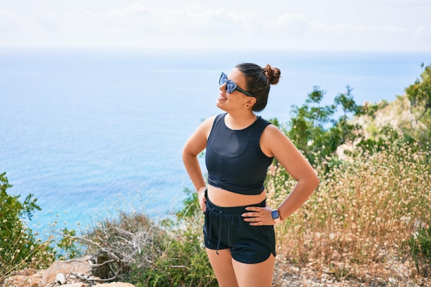Młoda latynoska kobieta ubrana w odzież sportową i okulary przeciwsłoneczne uśmiecha się szczęśliwa Stojąc z uśmiechem na twarzy nad pięknym krajobrazem