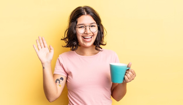 Młoda latynoska kobieta uśmiecha się radośnie, machając ręką, witając cię i pozdrawiając. koncepcja kubek kawy
