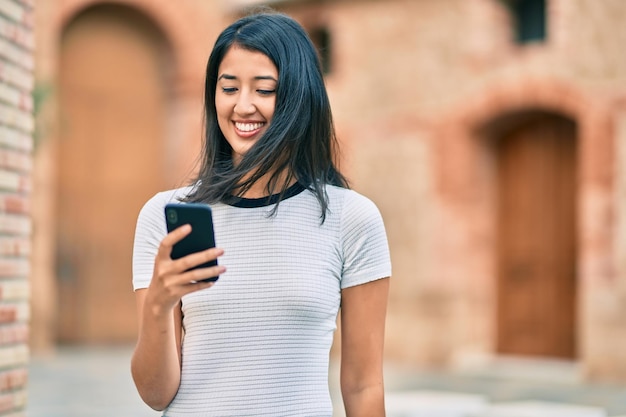 Młoda latynoska kobieta uśmiecha się szczęśliwa za pomocą smartfona w mieście.