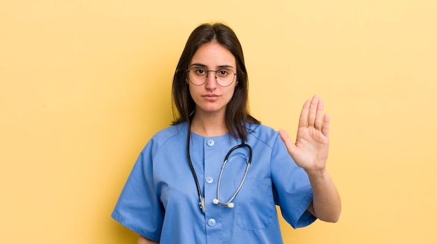 Młoda latynoska kobieta wygląda poważnie pokazując otwartą dłoń, robiąc koncepcję pielęgniarki gest zatrzymania