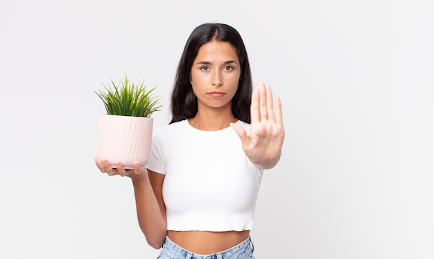 Młoda latynoska kobieta wygląda poważnie pokazując otwartą dłoń, wykonując gest zatrzymania i trzymając ozdobną roślinę domową