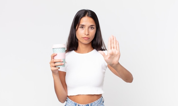 Młoda latynoska kobieta wygląda poważnie pokazując otwartą dłoń, wykonując gest zatrzymania i trzymający pojemnik na kawę na wynos