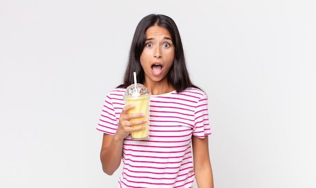 Młoda latynoska kobieta wyglądająca na bardzo zszokowaną lub zaskoczoną i trzymająca koktajl mleczny o smaku waniliowym