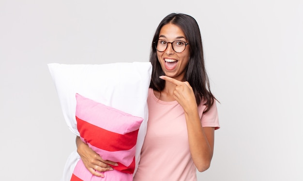 młoda latynoska kobieta wyglądająca na podekscytowaną i zaskoczoną, wskazując na bok, ubrana w piżamę i trzymająca poduszkę