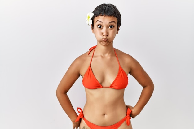 Młoda latynoska kobieta z krótkimi włosami ubrana w bikini nadymające policzki z zabawną buzią usta napompowane powietrzem szalonym wyrazem twarzy