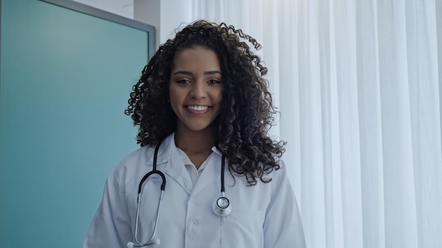 Młoda latynoska lekarka nosi biały mundur, biały fartuch medyczny, stetoskop i patrzy na kamerę