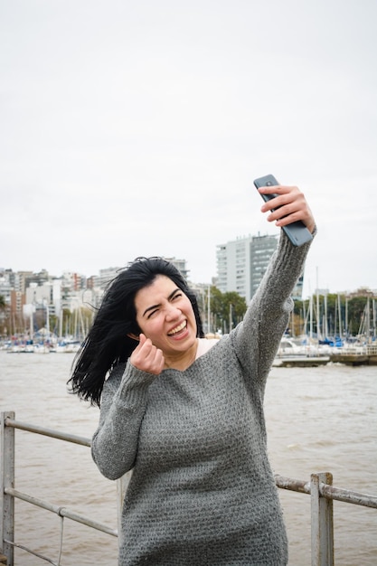 Młoda latynoska na nabrzeżu uśmiecha się szczęśliwie robiąc selfie z telefonem pionowe zdjęcie