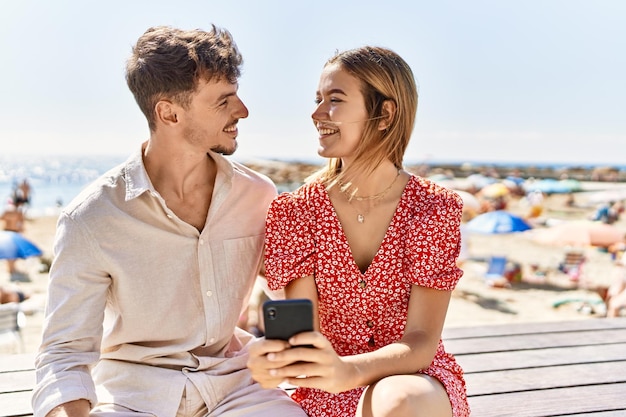 Młoda latynoska para na wakacjach za pomocą smartfona siedzącego na ławce na plaży.