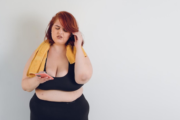 Młoda latynoska plus size z rudymi włosami i czarną odzieżą sportową za pomocą telefonu na białym tle