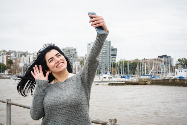 młoda latynoska turystka stojąca na molo robiąca sobie selfie ze swoim telefonem miasto na tle