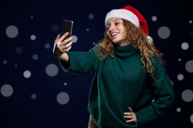 Młoda latynoska uśmiechnięta kobieta w swetrze i czapce Mikołaja robi selfie telefonem komórkowym