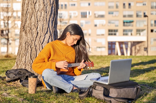 Młoda latynoska z długimi włosami grająca na ukulele pod drzewem ma laptopa, aby oglądać koncepcję muzyki leccion i uczyć się samouków