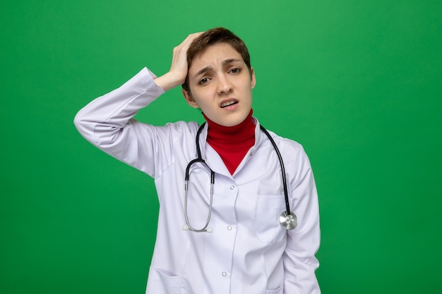 Młoda lekarka w białym fartuchu ze stetoskopem wygląda na zdezorientowaną, trzymając rękę na głowie za pomyłkę