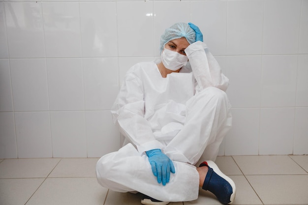 Zdjęcie młoda lekarka w masce ochronnej i rękawiczkach siedzi znużona na podłodze