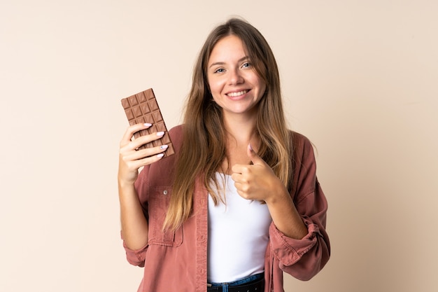 Młoda Litewska kobieta na białym tle na beżowym tle, biorąc czekoladową tabletkę iz kciukiem do góry