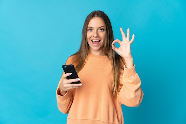 Młoda litewska kobieta na białym tle na niebieskiej ścianie przy użyciu telefonu komórkowego i robi znak OK