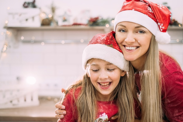 Młoda mama i nastoletnia córka w kuchni w czerwonej czapce Mikołaja śmieją się, a ich twarze są pokryte mąką