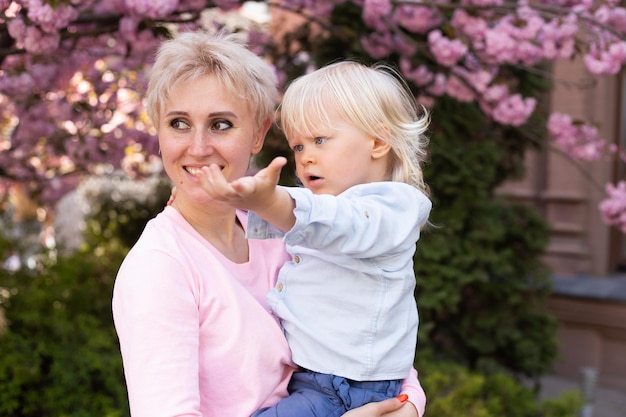 Młoda mama mama trzyma swojego małego synka pod kwitnącymi wiśniami Sakura z opadającymi różowymi płatkami i pięknymi kwiatami