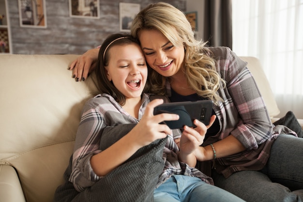Młoda Mama Pomaga Córeczce Z Szelkami Korzystać Z Aplikacji Ze Smartfona Siedzącego Na Kanapie W Ich Mieszkaniu.