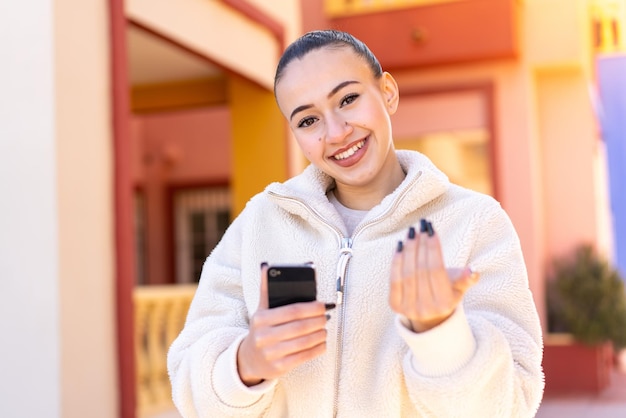 Młoda marokańska dziewczyna używająca telefonu komórkowego na zewnątrz zapraszająca do przyjścia z ręką Szczęśliwy, że przyszedłeś