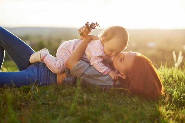 Młoda matka całuje małą córeczkę leżąc na trawie.
