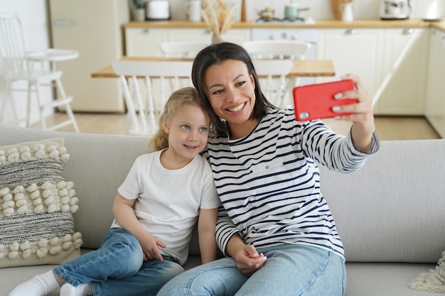 Młoda matka i adoptowana córka córeczki robią rodzinne zdjęcie selfie przez telefon, siedząc na kanapie w domu