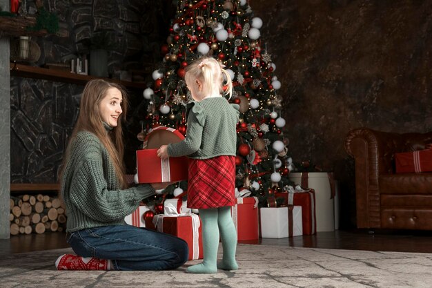 Młoda matka i jej mała córka otwierają prezent świąteczny dekoracja noworoczna w pomieszczeniach Mama i córka w pobliżu choiny w Wigilię