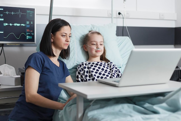Młoda matka i uśmiechnięta chora dziewczynka ogląda śmieszne filmy na laptopie. Matka i córka siedzi w pokoju na oddziale pacjenta kliniki pediatrycznej podczas korzystania z nowoczesnego komputera do rozrywki.