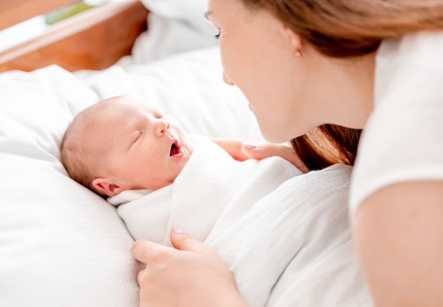 Młoda matka patrząca na swoją nowonarodzoną córkę owiniętą w białe prześcieradła w domu śpiącą z otwartymi ustami. Portret dziewczyny mamy z niemowlęciem drzemiącym w łóżku