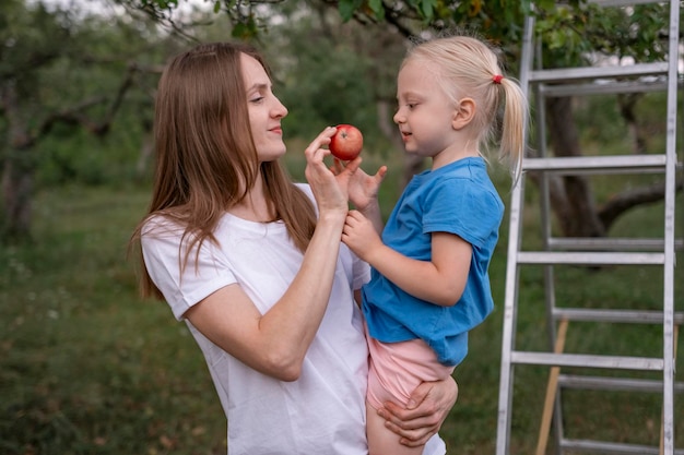 Młoda matka traktuje córkę blondynkę do dojrzałych czerwonych jabłek Mama i dziecko zbiera jabłka w ogrodzie