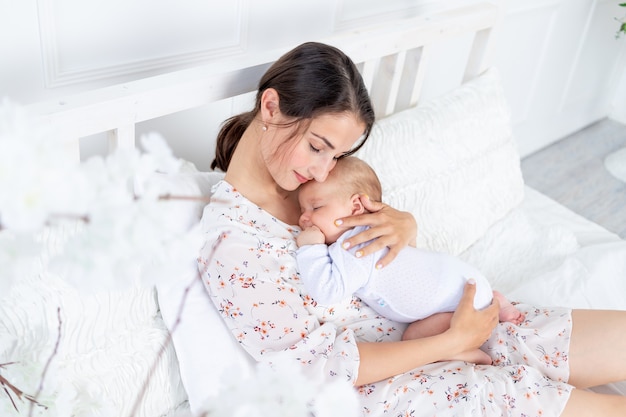 Młoda matka trzyma w ramionach śpiącego noworodka na łóżku w sypialni, pojęcie macierzyństwa i szczęśliwej rodziny.