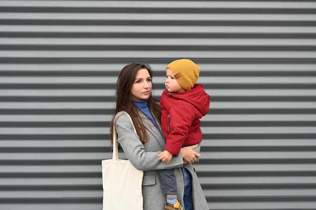 Młoda matka w szarym płaszczu trzyma chłopca w ramionach na tle szarej ściany
