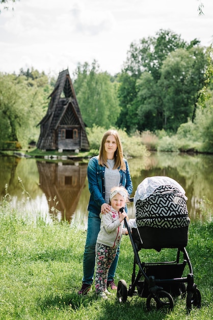 Młoda matka z dwójką dzieci i wózkiem spaceruje nad jeziorem w naturze Na tle starego drewnianego domu z mostem prowadzącym przez rzekę Spacery w parku