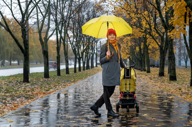 Młoda matka z wózkiem dziecięcym i żółtym parasolem spaceruje aleją jesiennego parku.