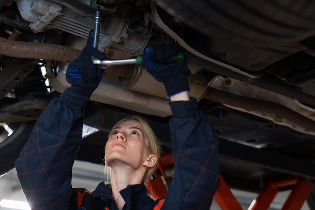 Zdjęcie młoda mechanik samochodowy odkręca śruby w zawieszeniu samochodu, który jest na windzie na tle centrum serwisowego