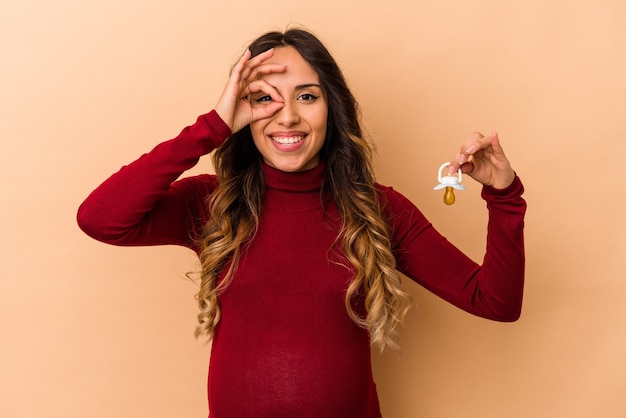 Młoda meksykańska kobieta w ciąży trzymając smoczek na białym tle na beżowym tle podekscytowany, trzymając ok gest na oko.