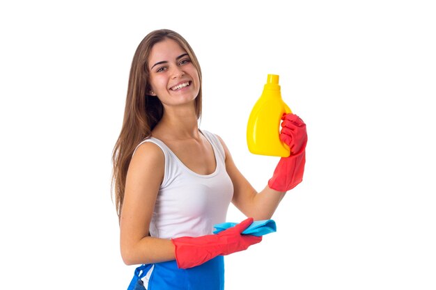 Młoda miła kobieta w białej koszuli i niebieskim fartuchu z czerwonymi rękawiczkami, trzymająca szmatkę i detergent