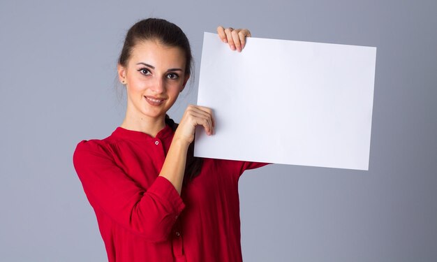 Młoda miła kobieta w bluzce z warkoczem, trzymająca białą kartkę papieru na szarym tle w studio