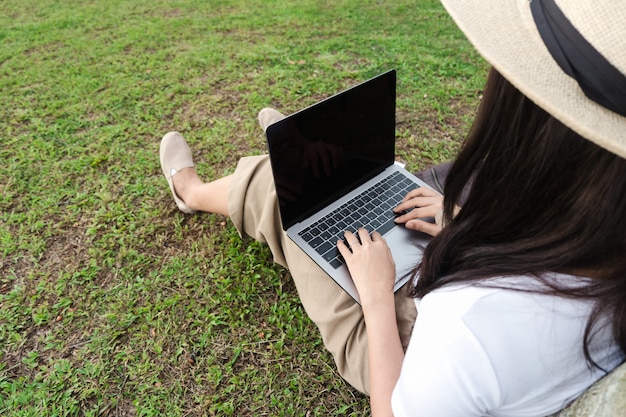 Zdjęcie młoda modniś kobieta używa laptop podczas gdy siedzący na trawie z torbą i pustym notatnikiem.