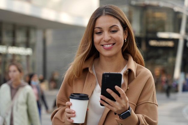 Młoda multi etniczna kobieta biznesu spaceruje oglądając telefon komórkowy z zamazanymi ludźmi na współczesnym tle miejskim