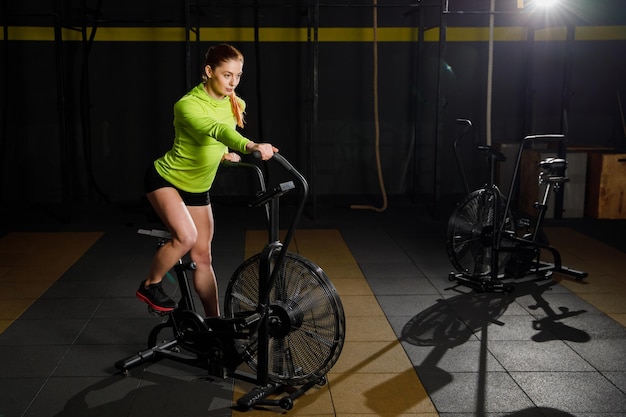 Młoda muskularna kobieta ćwiczy crossfit na rowerze w siłowni.