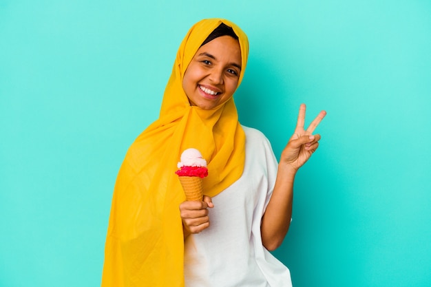 Młoda muzułmanka jedząca lody na białym tle na niebieskiej ścianie radosna i beztroska pokazująca palcami symbol pokoju.