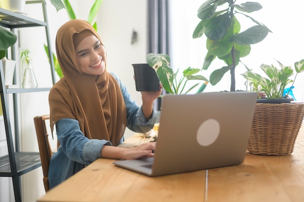 Młoda muzułmanka, przedsiębiorca pracująca z laptopem, prezentuje rośliny doniczkowe podczas transmisji online na żywo w domu, sprzedając koncepcję online