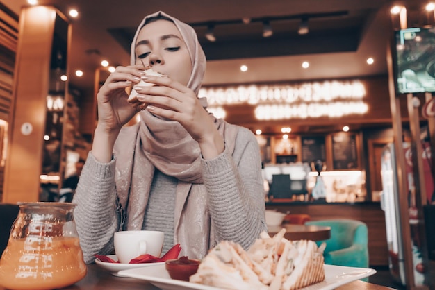 Młoda muzułmanka w chuście na głowie je w przytulnej restauracji pyszne jedzenie i pachnącą herbatę