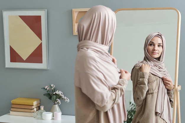 Młoda muzułmanka w hidżabie patrząca w duże lustro podczas ubierania się rano do pracy