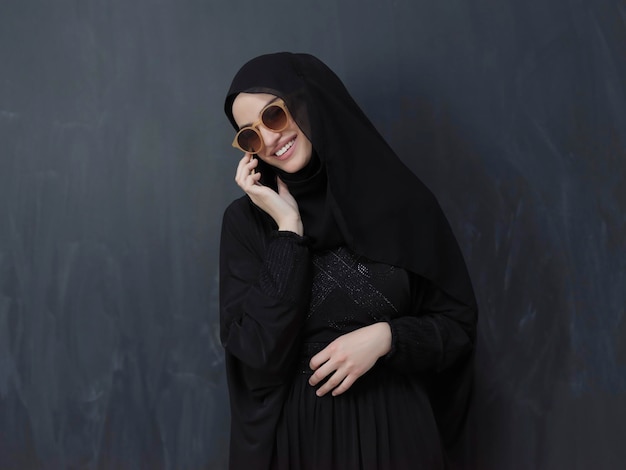 Młoda muzułmańska bizneswoman w tradycyjnych strojach lub abaya za pomocą smartfona. Arabska kobieta szczęśliwa stojąc przed czarną tablicą i reprezentująca technologię, islamską modę i Ramadan karee