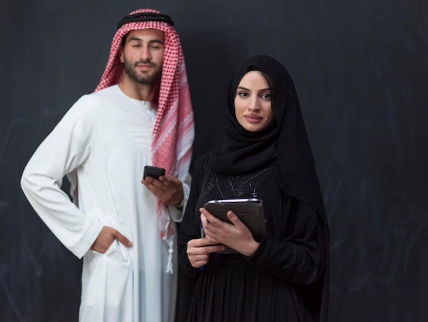 Młoda muzułmańska para biznesowa w modnej sukience hidżabu za pomocą smartfona i tabletu przed czarnym tłem. Wysokiej jakości zdjęcie